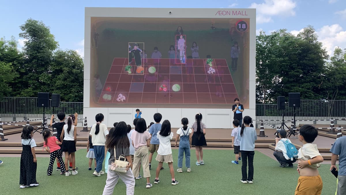 ショッピングモールの屋外の大型スクリーンの前で、体を動かしてARゲームを遊ぶ子どもたち